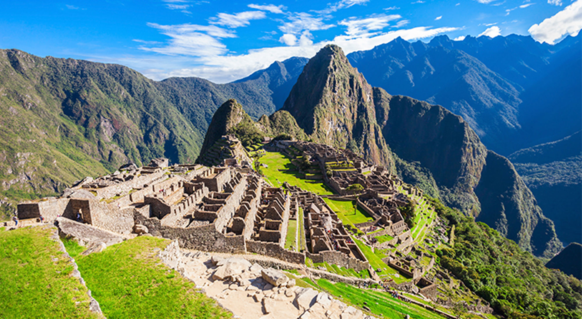 Panoramisch uitzicht op Machu Picchu met groene terrassen en bergen onder een blauwe lucht.