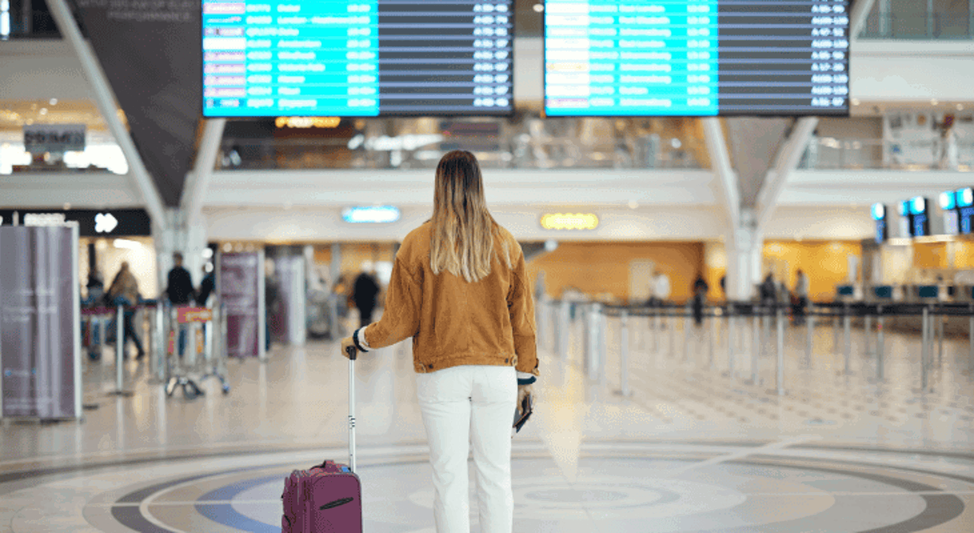 Luchthaven Reiziger met rolkoffer in luchthavenhal, kijkend naar vluchtinformatie op vertrekborden.
