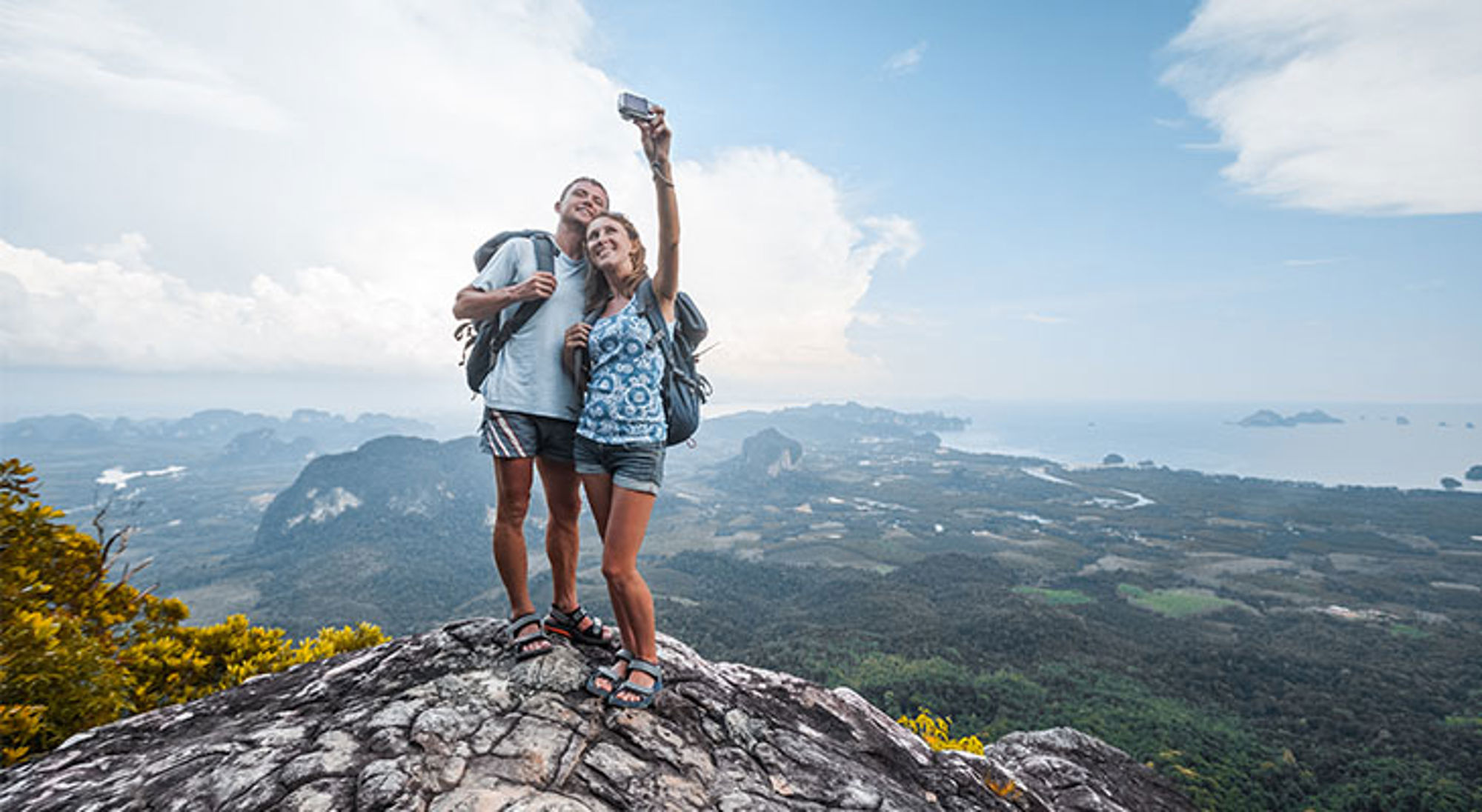 Wandelaars met rugzak maken een selfie op een bergtop met uitzicht over groene valleien en zee.