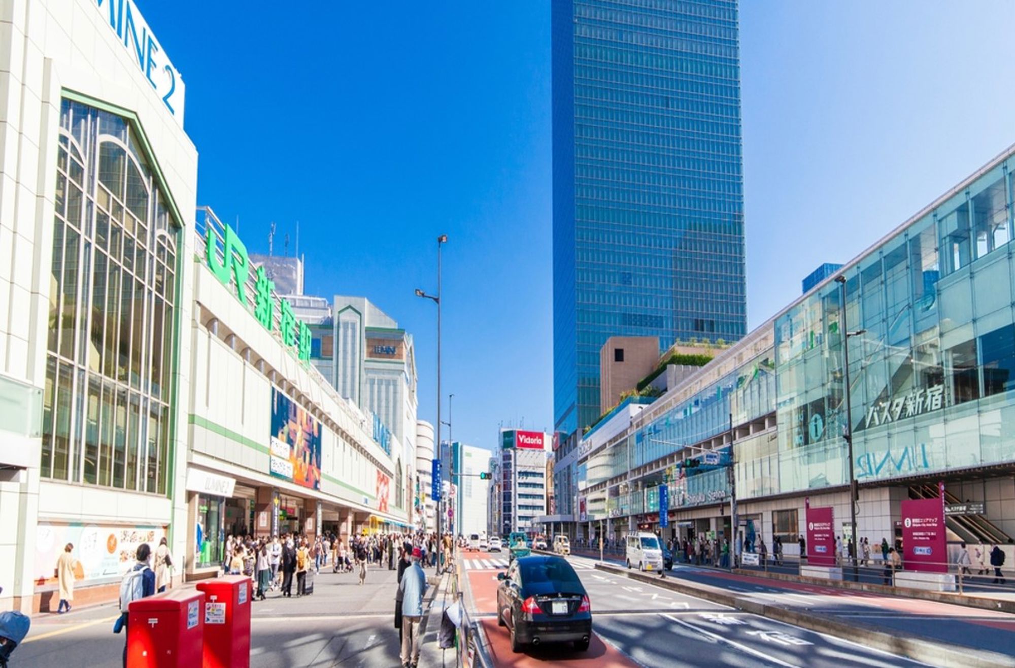 Drukke straat bij station Shinjuku in Tokio met hoge gebouwen, winkels en voetgangers op een zonnige dag.