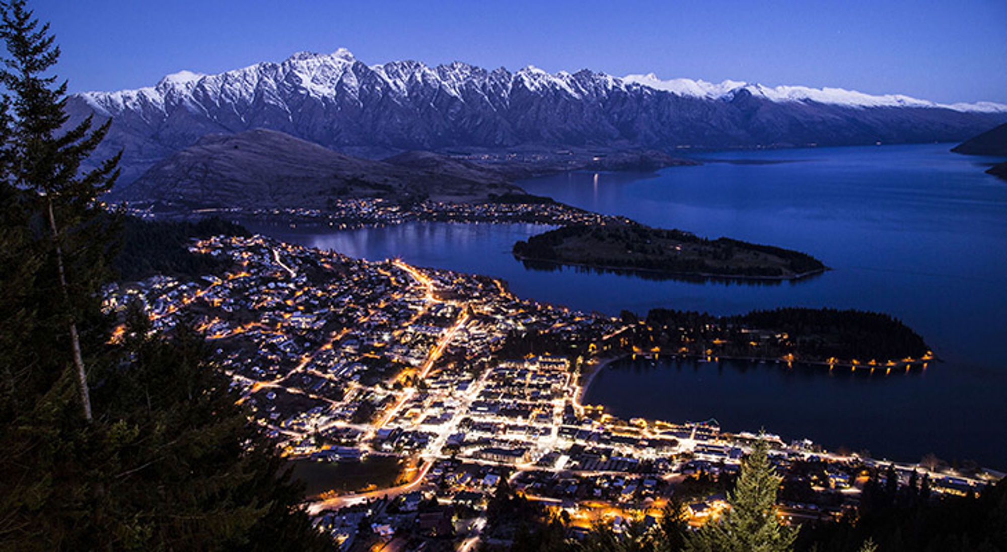 nieuw-zeeland-queenstown Avondpanorama van Queenstown aan Lake Wakatipu met besneeuwde bergen op de achtergrond.