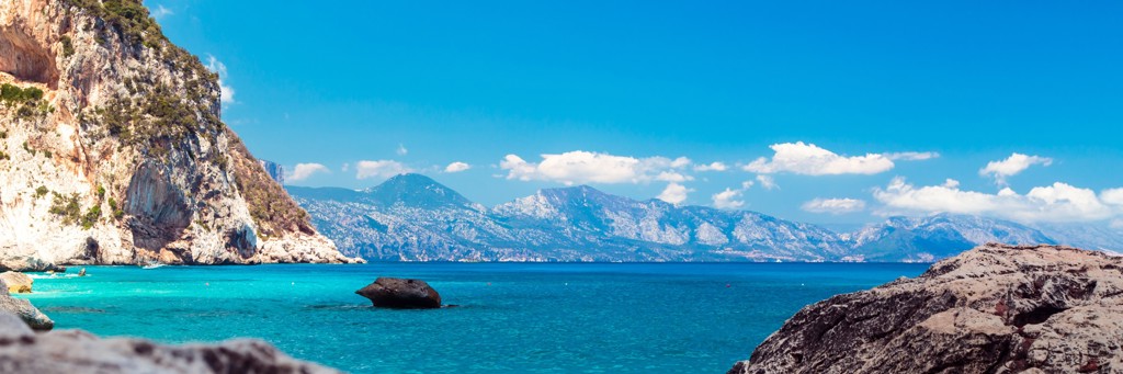 Rotsachtige kust en turquoise zee op Sardinië, Italië, met bergen op de achtergrond.