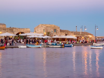 Avond in een vissersdorp op Sicilië met terrasjes aan de haven en kleurrijke bootjes op het water.
