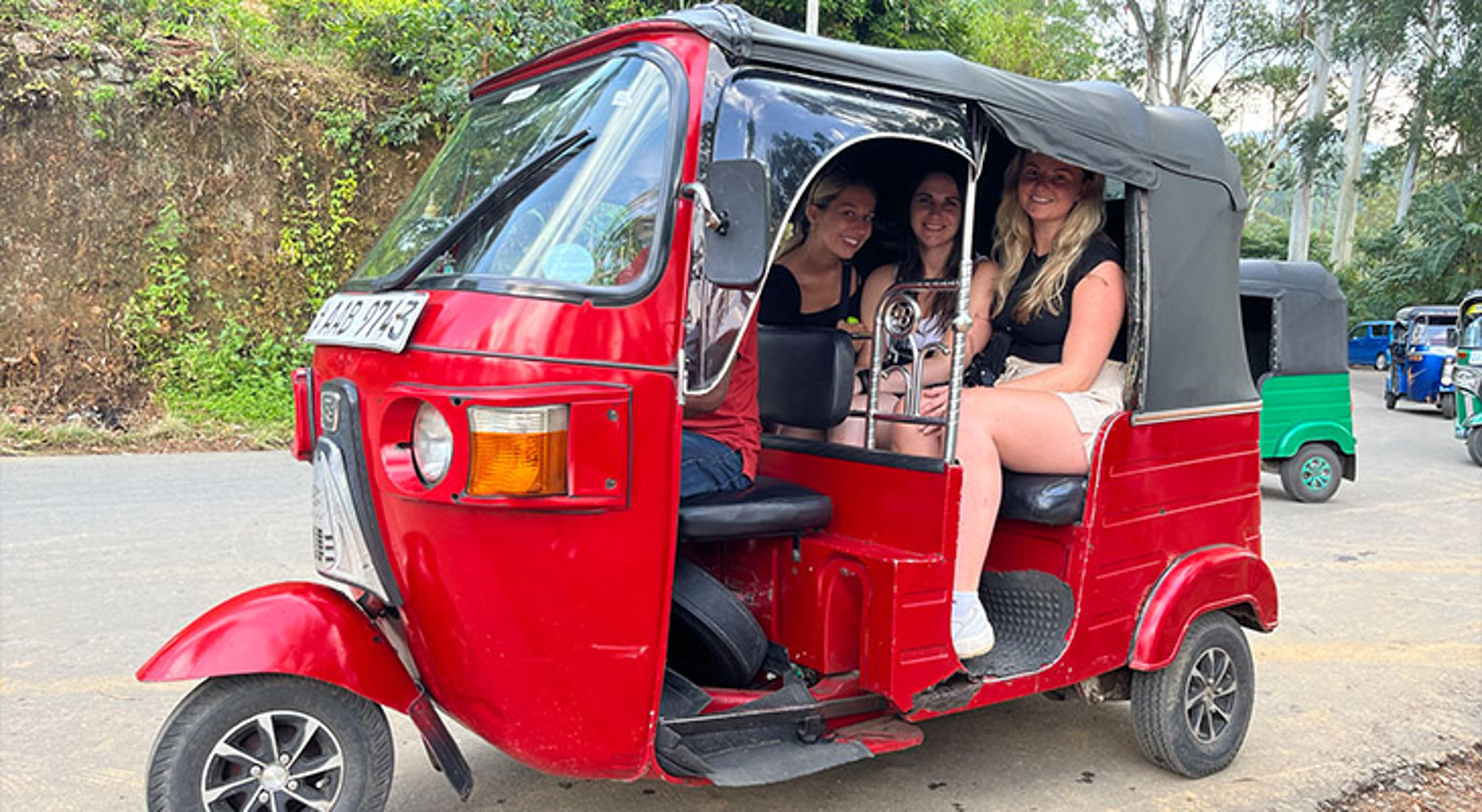 Vrouwen in een rode tuk-tuk tijdens een rit over een weg met groen landschap op de achtergrond.