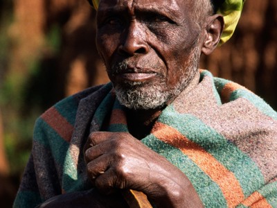 Portret van een man in Ethiopië met kleurrijke hoofddoek en omslagdoek, leunend op een wandelstok.