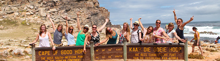 Jongvolwassenen op groepsreis poseren bij het bord Cape of Good Hope aan de kust.
