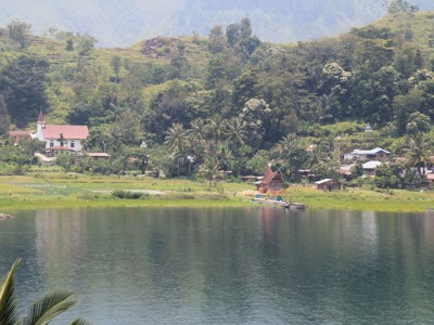 Dorp aan de oever van het Tobameer op Sumatra, met groene heuvels en palmbomen op de achtergrond.