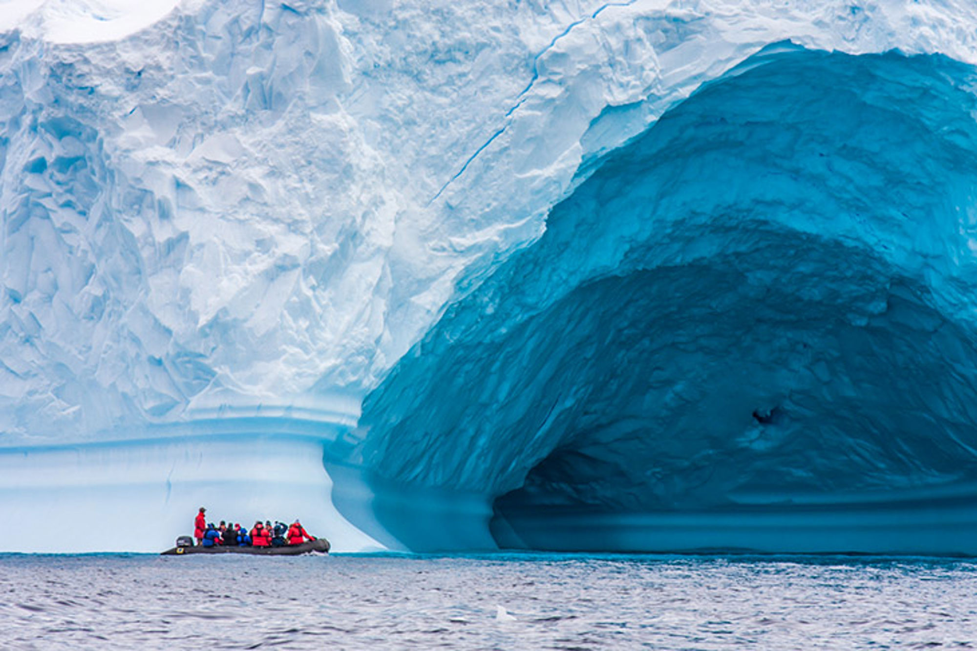 Bootje Antarctica Zodiacboot met reizigers langs een enorme ijsberg met blauwe ijsgrot op zee.