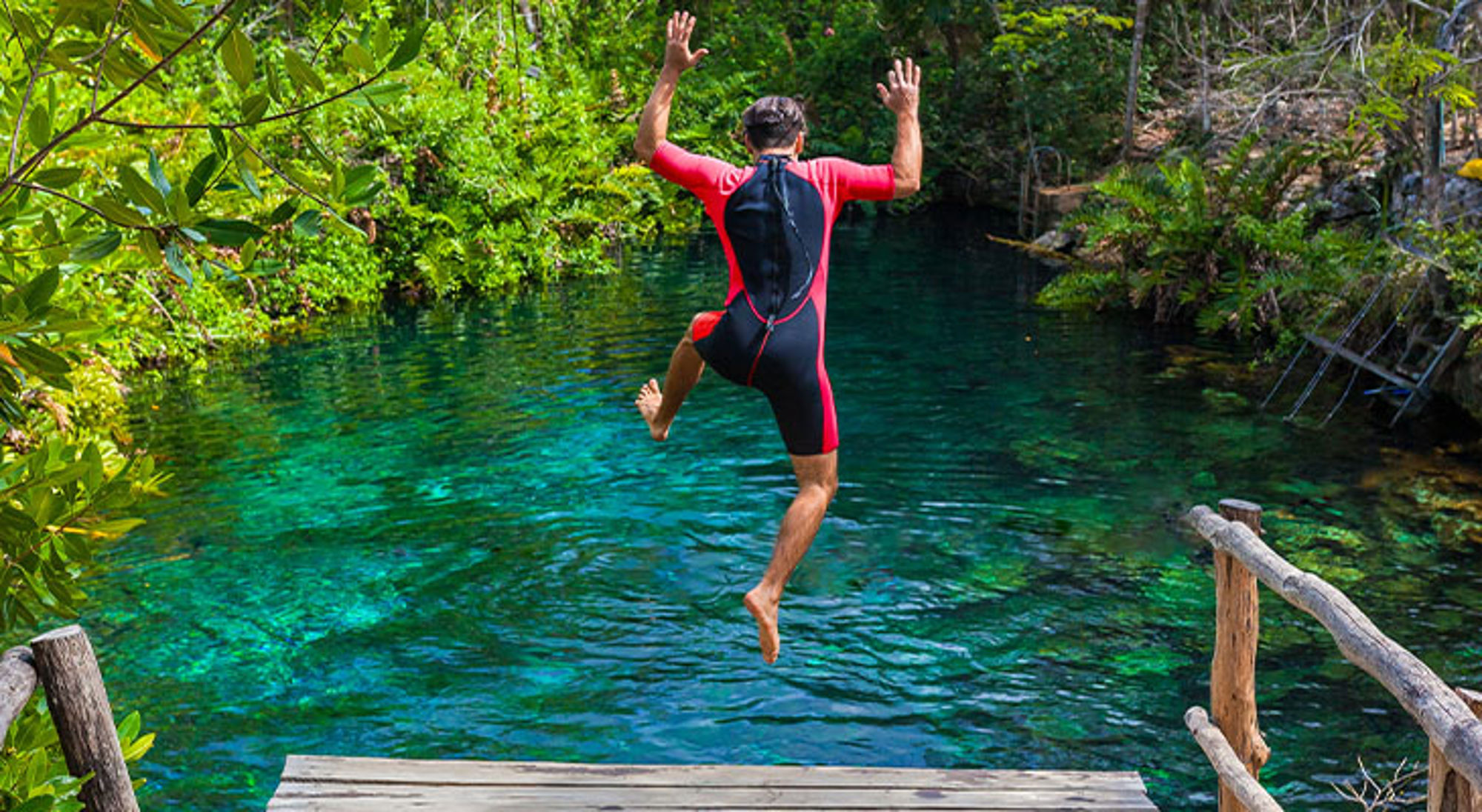 Cenote Mexico Man springt vanaf een houten steiger in een helderblauwe natuurlijke zwemvijver in het groen.