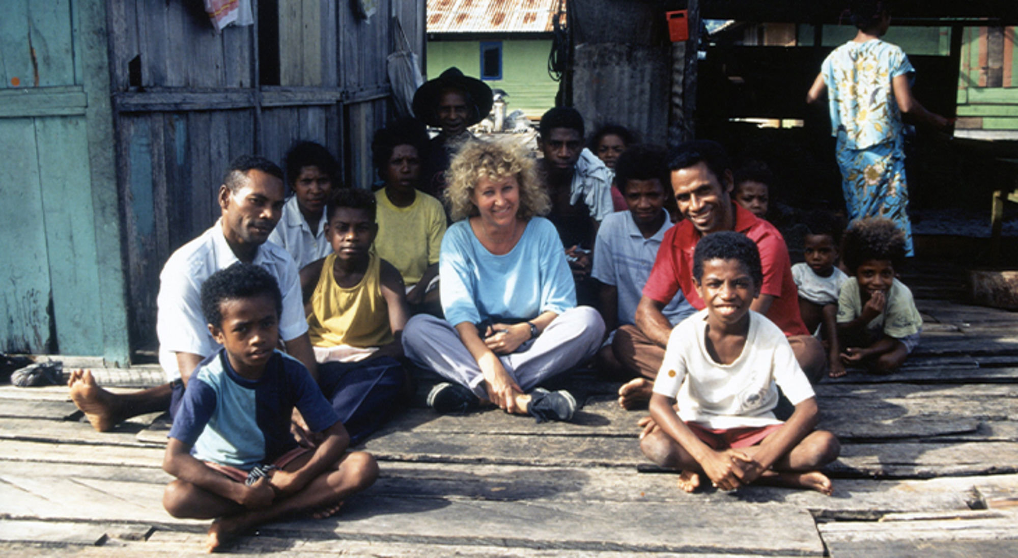 Mieke in Papua Groepsportret van lokale bewoners en een reiziger op een houten veranda bij eenvoudige huizen.