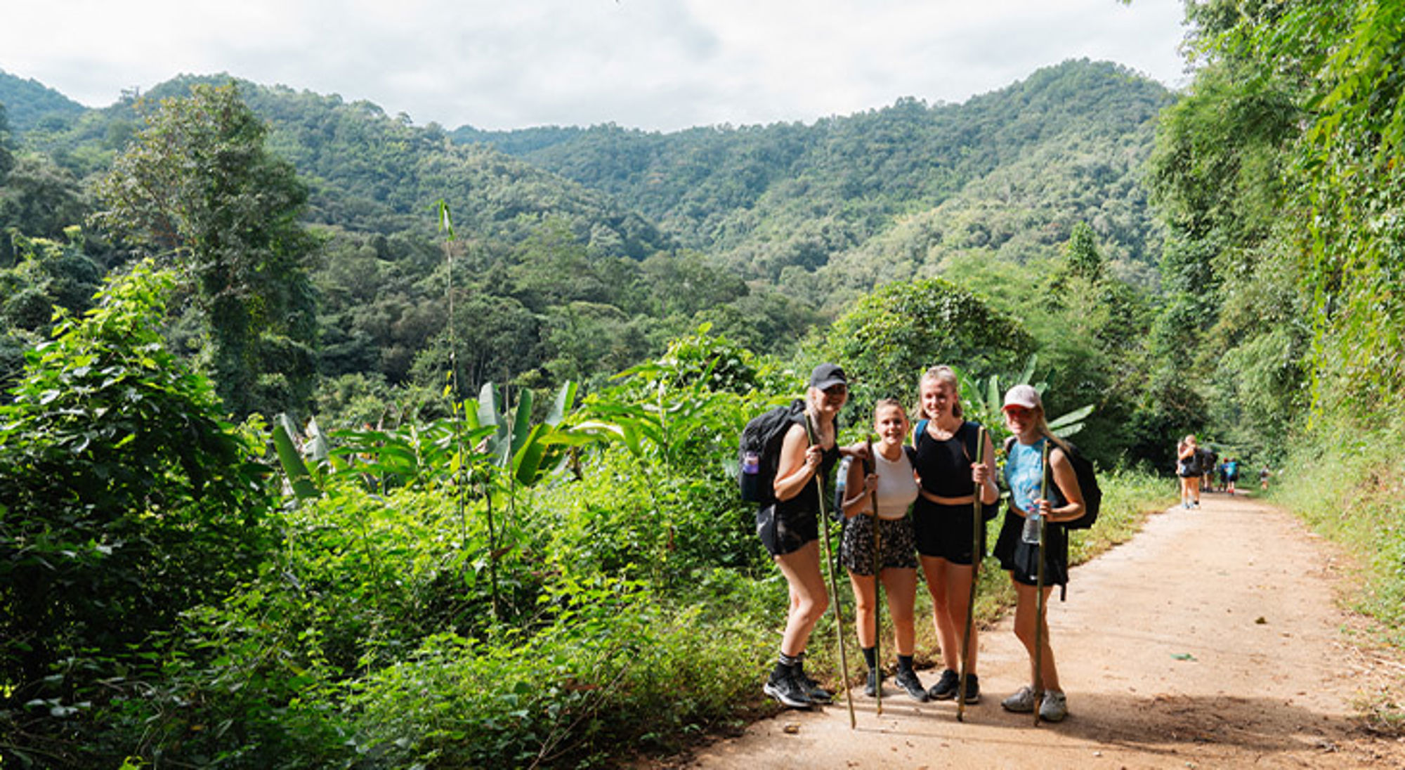 Vier wandelaars met rugzakken poseren op een pad in een groene, bergachtige jungle.