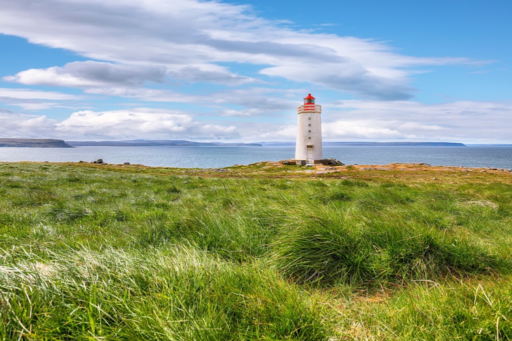 Vuurtoren aan de kust van IJsland, met grasland op de voorgrond en zee onder een blauwe lucht.