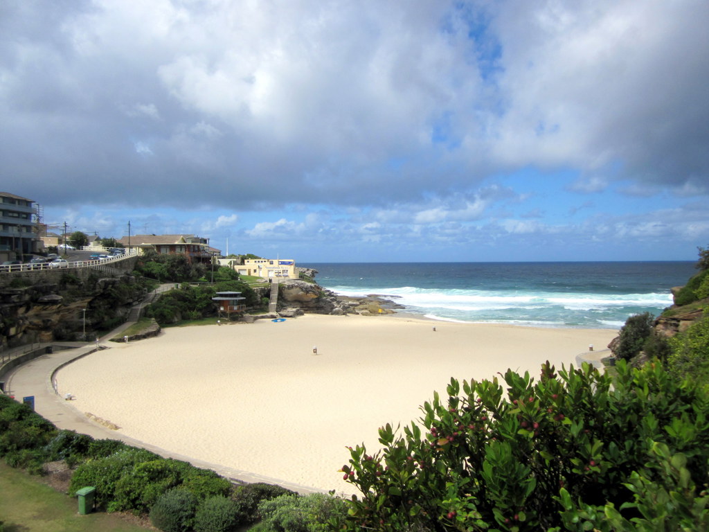 Zandstrand en branding bij een baai aan de kust van Australië, met wandelpad en bebouwing op de kliffen.