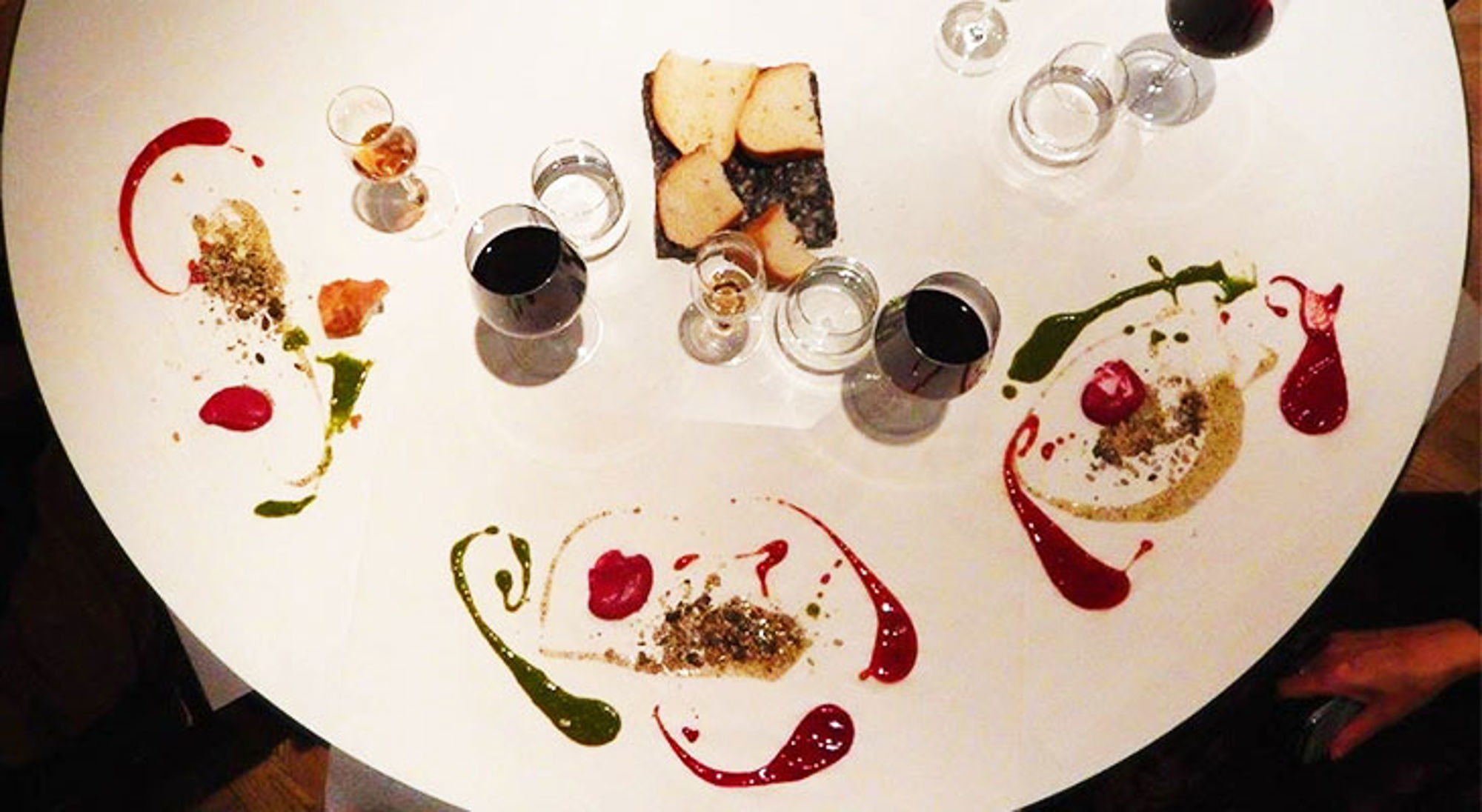 Gedekte tafel met borden vol kleurrijke sausdecoraties, brood en glazen rode wijn.