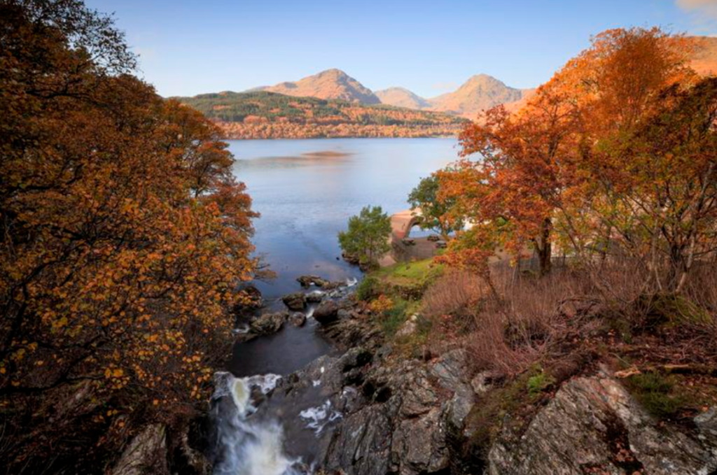 Herfstlandschap bij een loch in de Schotse Hooglanden met waterval, bergen en gekleurde bomen.