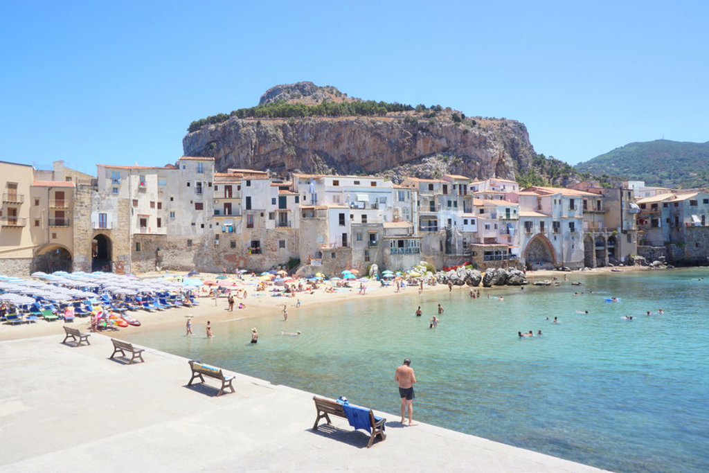 Strand en kustplaats Cefalù op Sicilië met kleurrijke parasols en historische huizen aan zee.