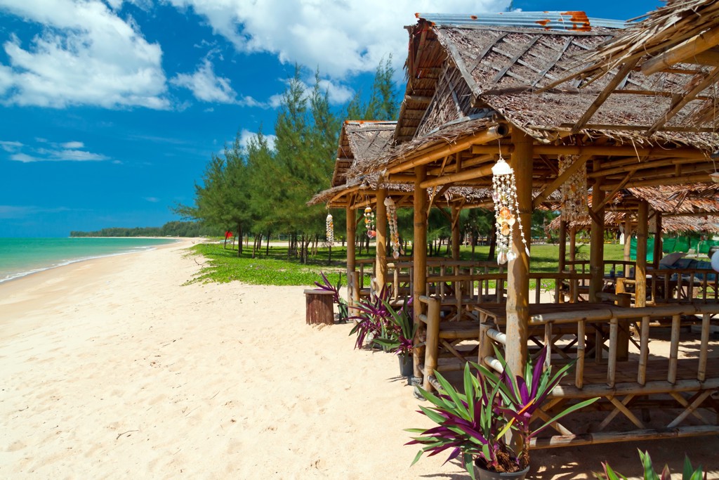 Bamboehutjes aan een wit zandstrand in Zuid-Thailand, met zee, pijnbomen en blauwe lucht.