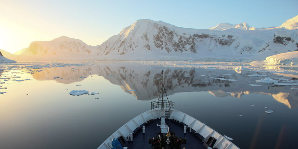 Expeditiecruise schip vaart tussen ijsbergen langs besneeuwde bergen in Antarctica bij zonsopkomst.