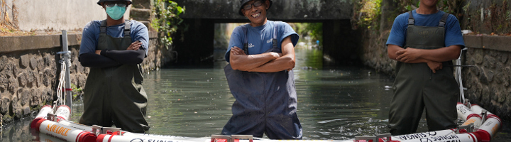 Sungai Watch-team in waadpakken in een waterkanaal met drijvende afvalbarrière.