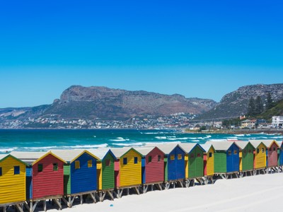 Kleurrijke strandhuisjes aan Muizenberg Beach bij Kaapstad, met zee en bergen op de achtergrond.
