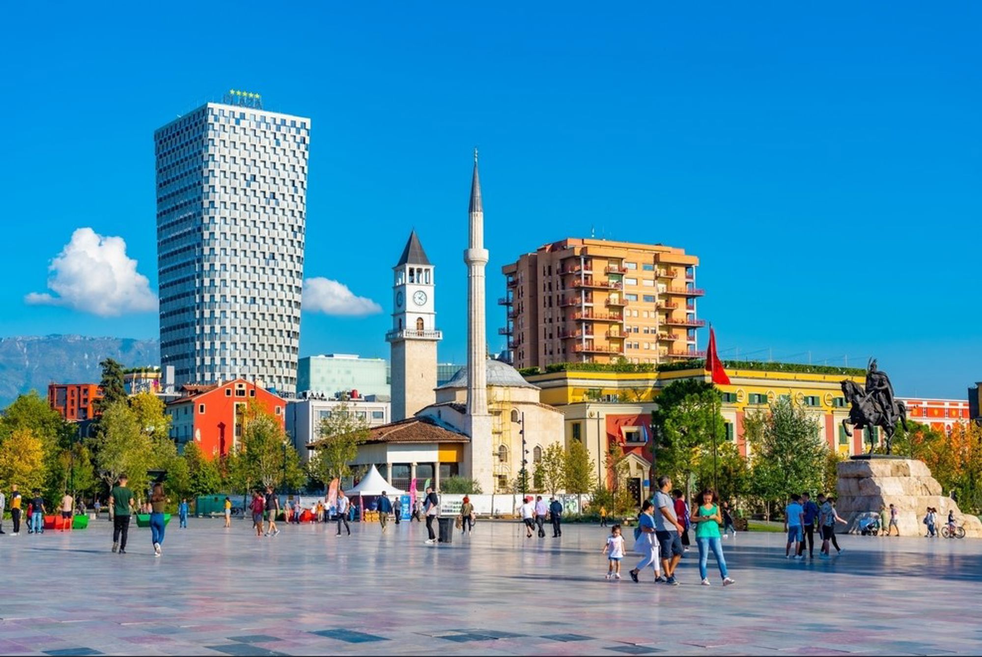 Skanderbegplein in Tirana met Et'hem Bey-moskee, klokkentoren en moderne skyline onder blauwe lucht.