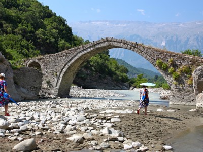 Oude stenen boogbrug over een rivier in Albanië, met bergen op de achtergrond.