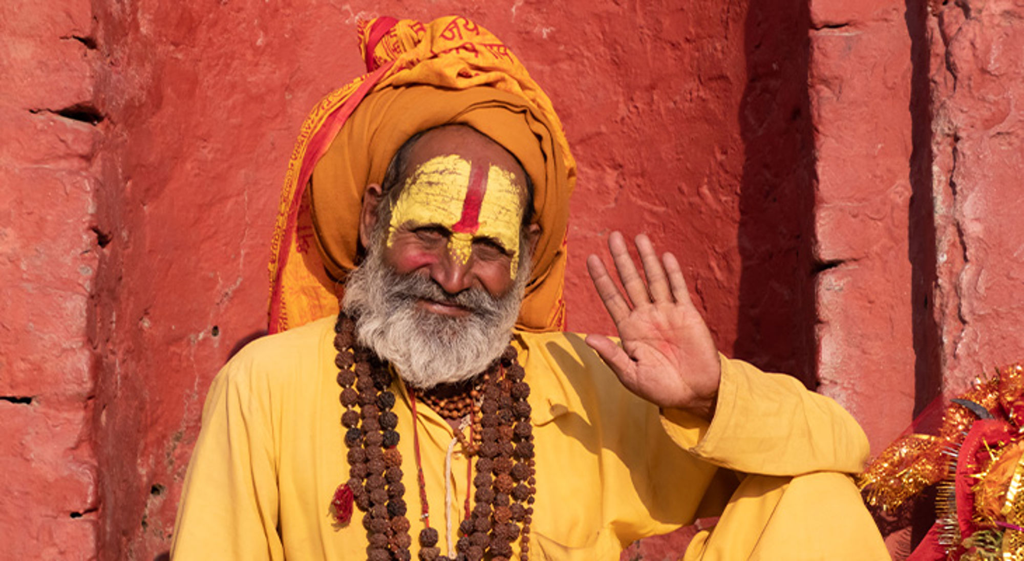 Vraag toestemming voordat je een local fotografeert Portret van een hindoe sadhu in oranje gewaad met gezichtsbeschildering, zittend tegen een rode muur en zwaaiend.