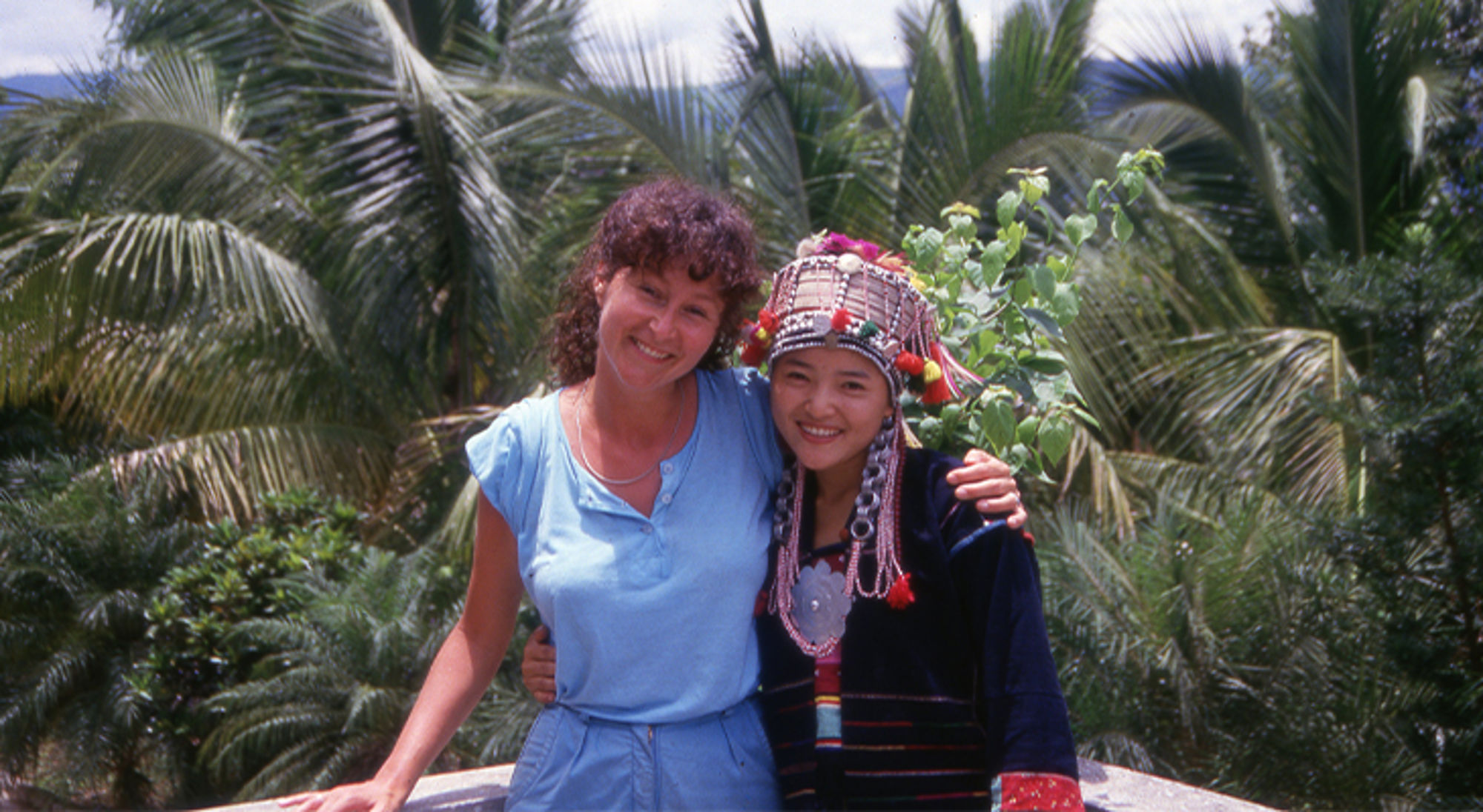 Mieke in China Twee vrouwen poseren en glimlachen tussen palmbomen, één draagt traditionele kleding met sierlijke hoofdtooi.