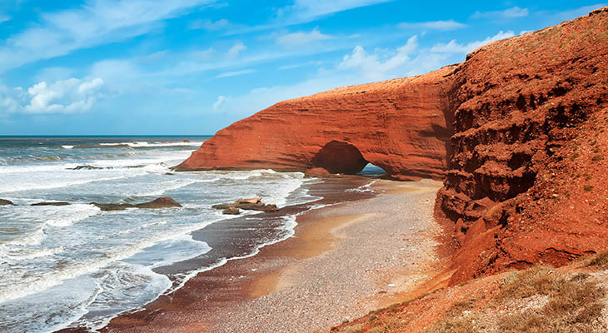 Legzira Beach Rode rotsboog aan de Atlantische kust van Marokko, met golven en zandstrand onder een blauwe lucht.