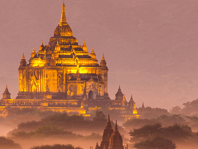 Gouden tempels en pagodes in Bagan bij zonsopkomst, met nevel en bergen op de achtergrond.