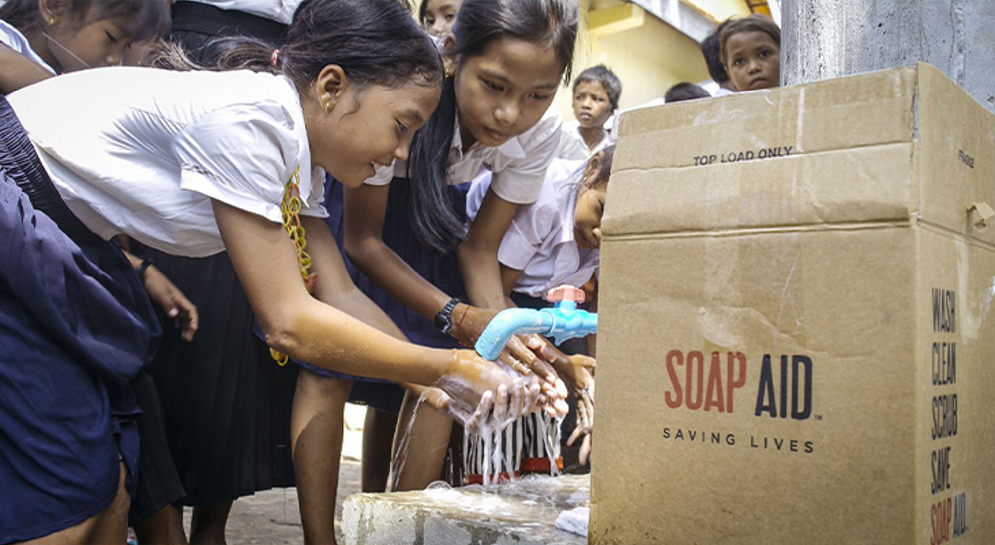 Donatie Soap Aid Schoolkinderen wassen hun handen bij een kraan met zeep, naast een doos met “SOAP AID”.
