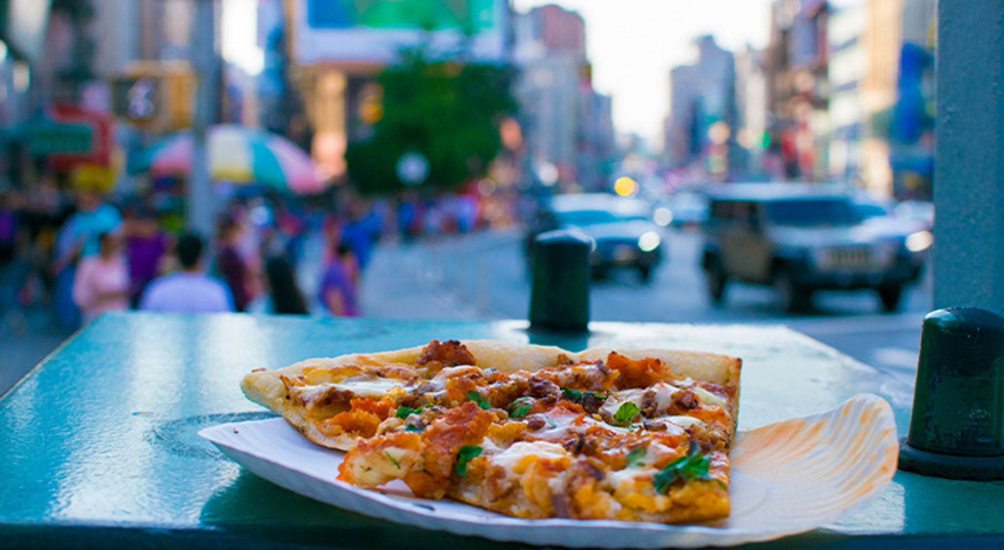 Streetfood New York Punt pizza op een papieren bordje op straat, met druk stadsverkeer op de achtergrond.