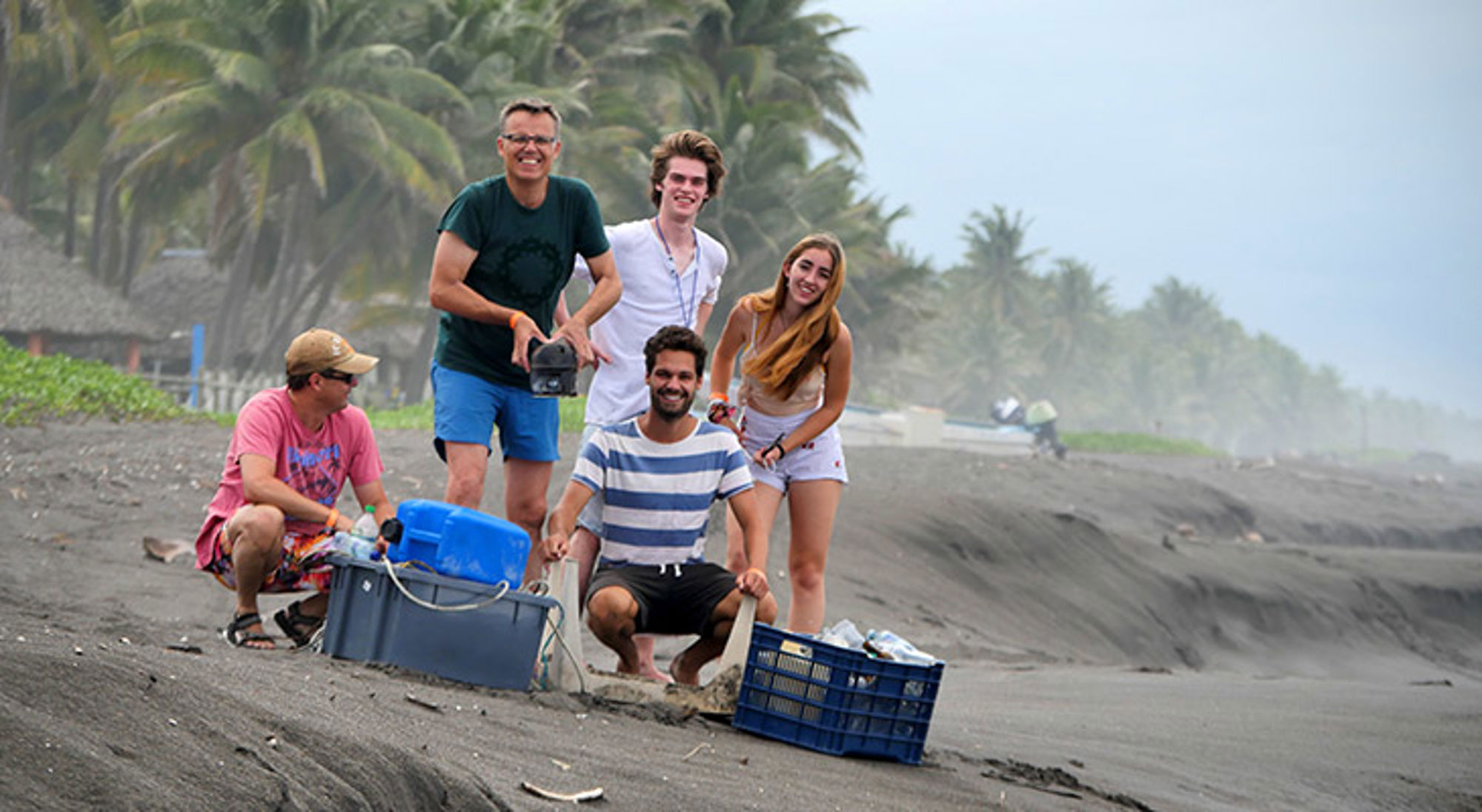 Groep reizigers op een zwart zandstrand met palmbomen, bij bagage en uitrusting.