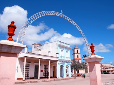 Pastelkleurige gevels en boogvormige toegangspoort op een plein in Cuba, met kerktoren op de achtergrond.