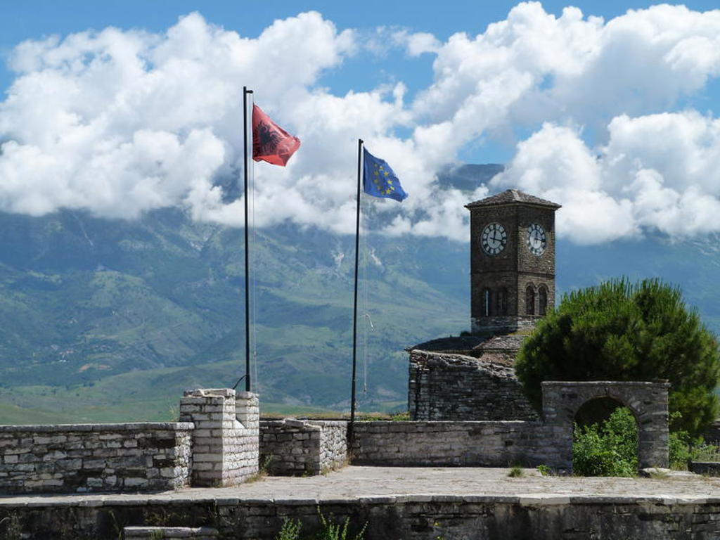 Klokkentoren met Albanese en EU-vlag bij kasteel, met bergen en wolken op de achtergrond.