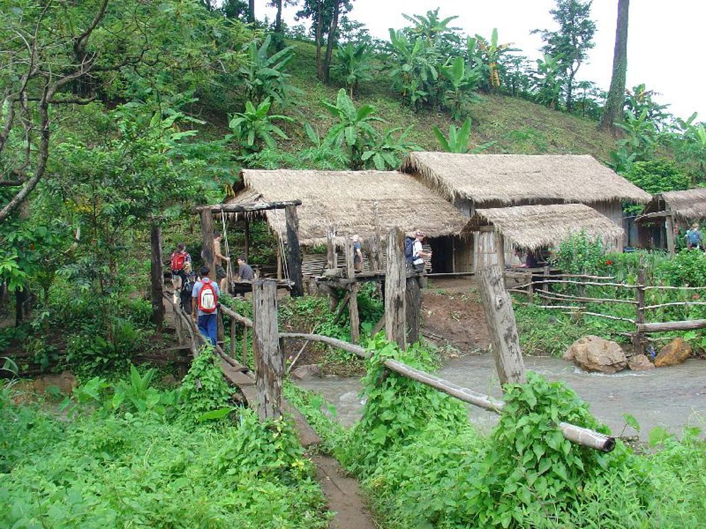 Gezin wandelt over een houten brug naar een bergdorp met rieten huizen in het groene noorden van Thailand.