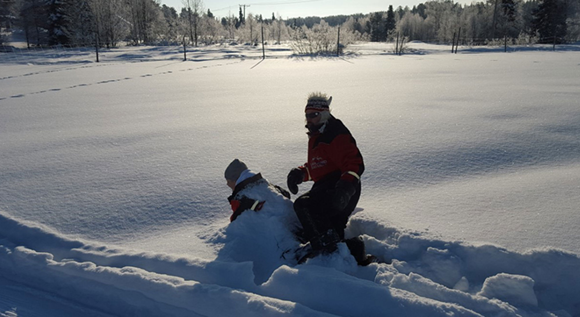 Man in winterkleding maakt een sneeuwpop in een besneeuwd landschap met bomen op de achtergrond.