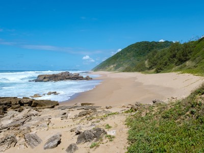 Zandstrand aan de Wild Coast in Zuid-Afrika met rotsen, golven en groene duinen onder een blauwe lucht.