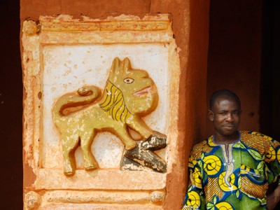 Man in kleurrijke outfit naast reliëf van een leeuw op een roodbruine muur in Ghana, Togo of Benin.