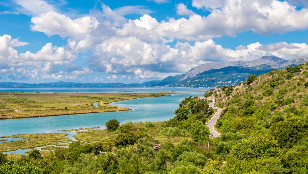 Kronkelende kustweg langs een blauwe baai met bergen op de achtergrond in Albanië.