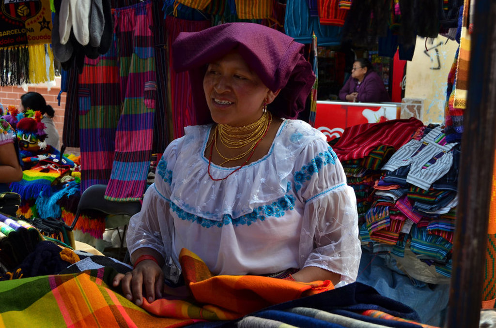 Marktvrouw in traditionele kleding tussen kleurrijke textielkramen tijdens een groepsrondreis door Ecuador.