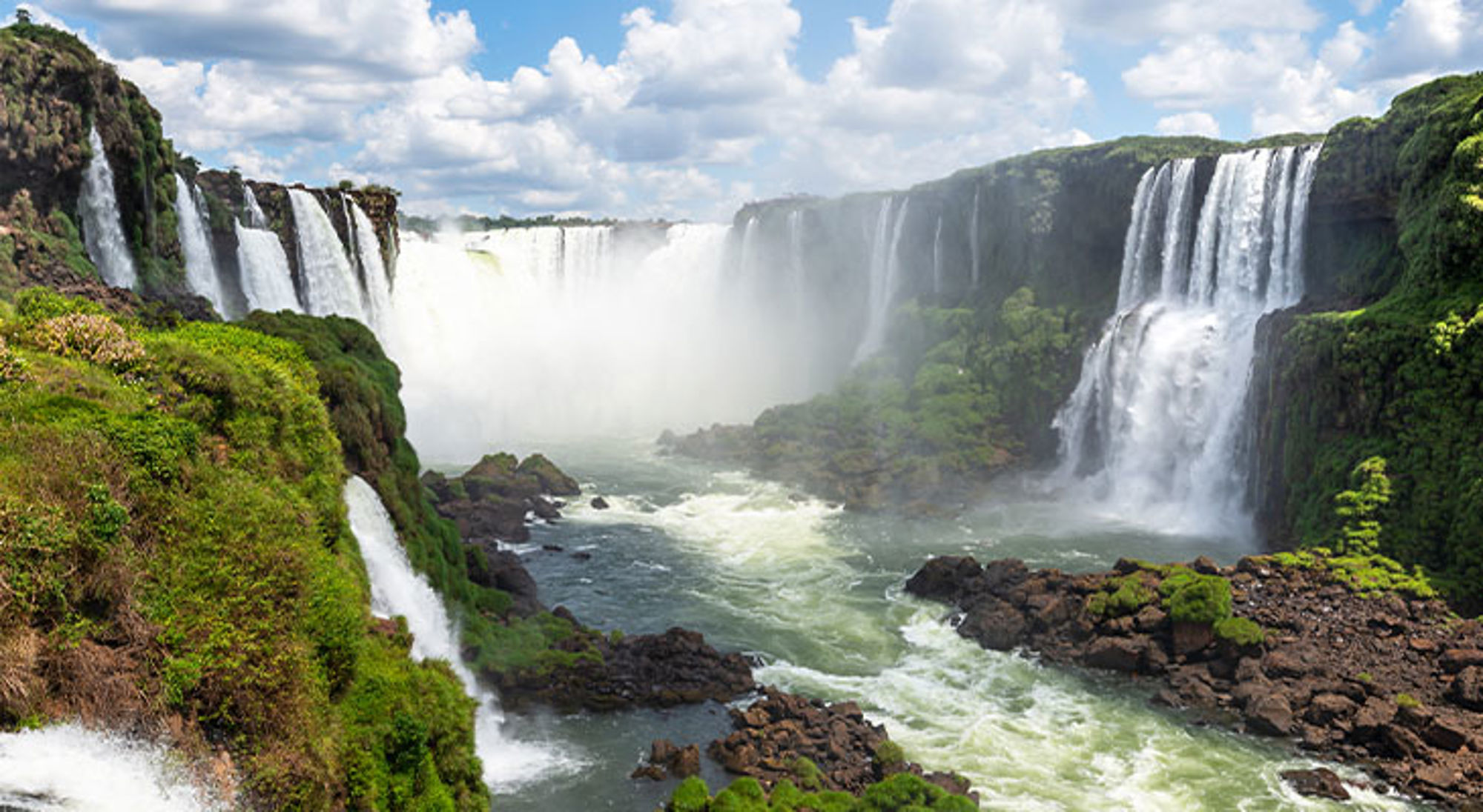 Iguazu Falls - Argentinië & Brazilië Panorama van de Iguazú-watervallen met meerdere watervallen, groene rotswanden en opspattende nevel onder een blauwe lucht.