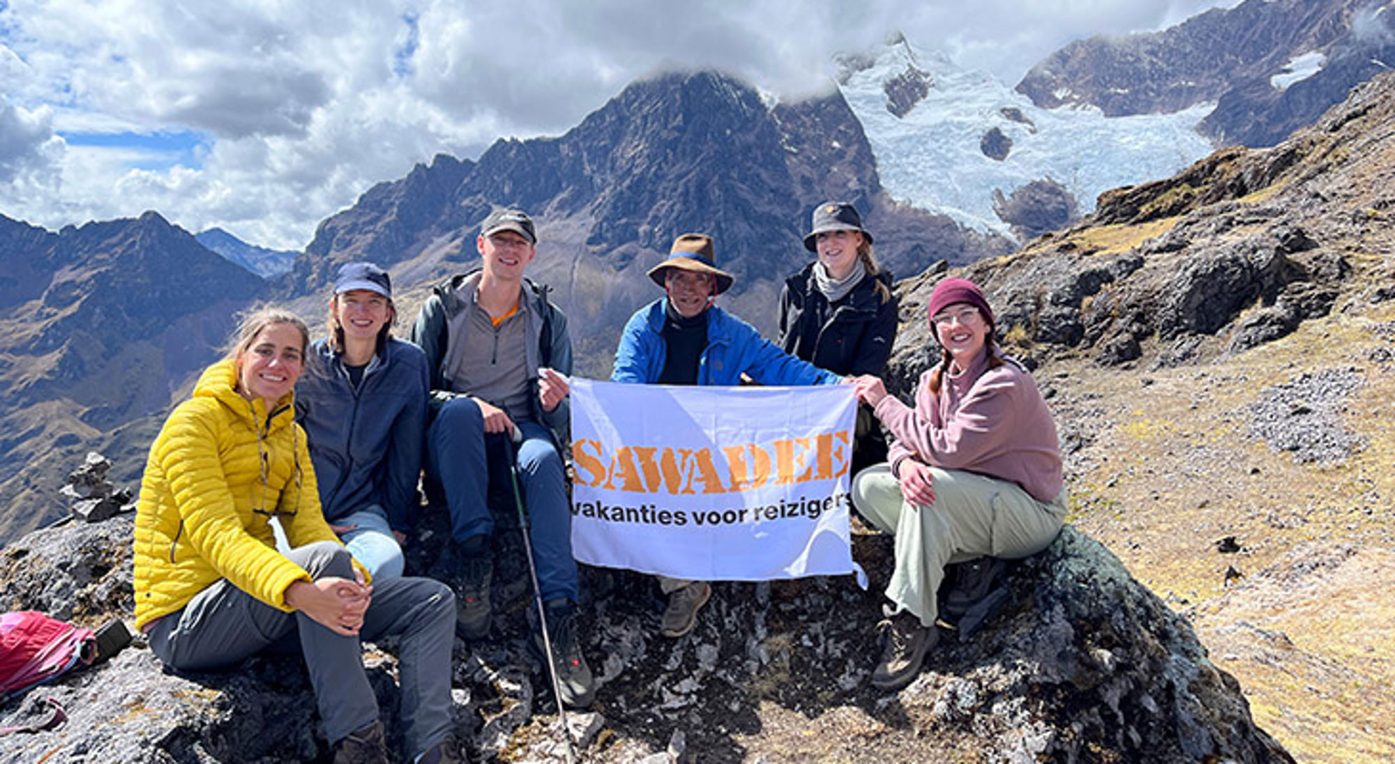 Larestrail Groep wandelaars met Sawadee-banner op bergtop met gletsjer en ruige bergketen op de achtergrond.