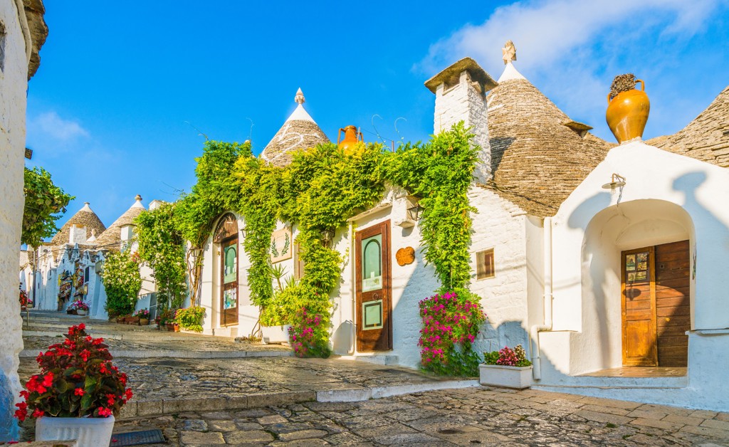 Trulli-huizen met kegelvormige daken in Alberobello, Puglia, langs een geplaveide straat met bloemen.