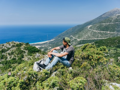Uitzicht op de Albanese Rivièra tijdens de Route Albanië, met reiziger op rots en bergen boven de zee.