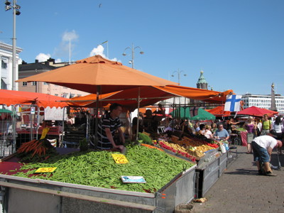 Drukke markt met groentestallen onder oranje parasols en een Finse vlag op het plein.