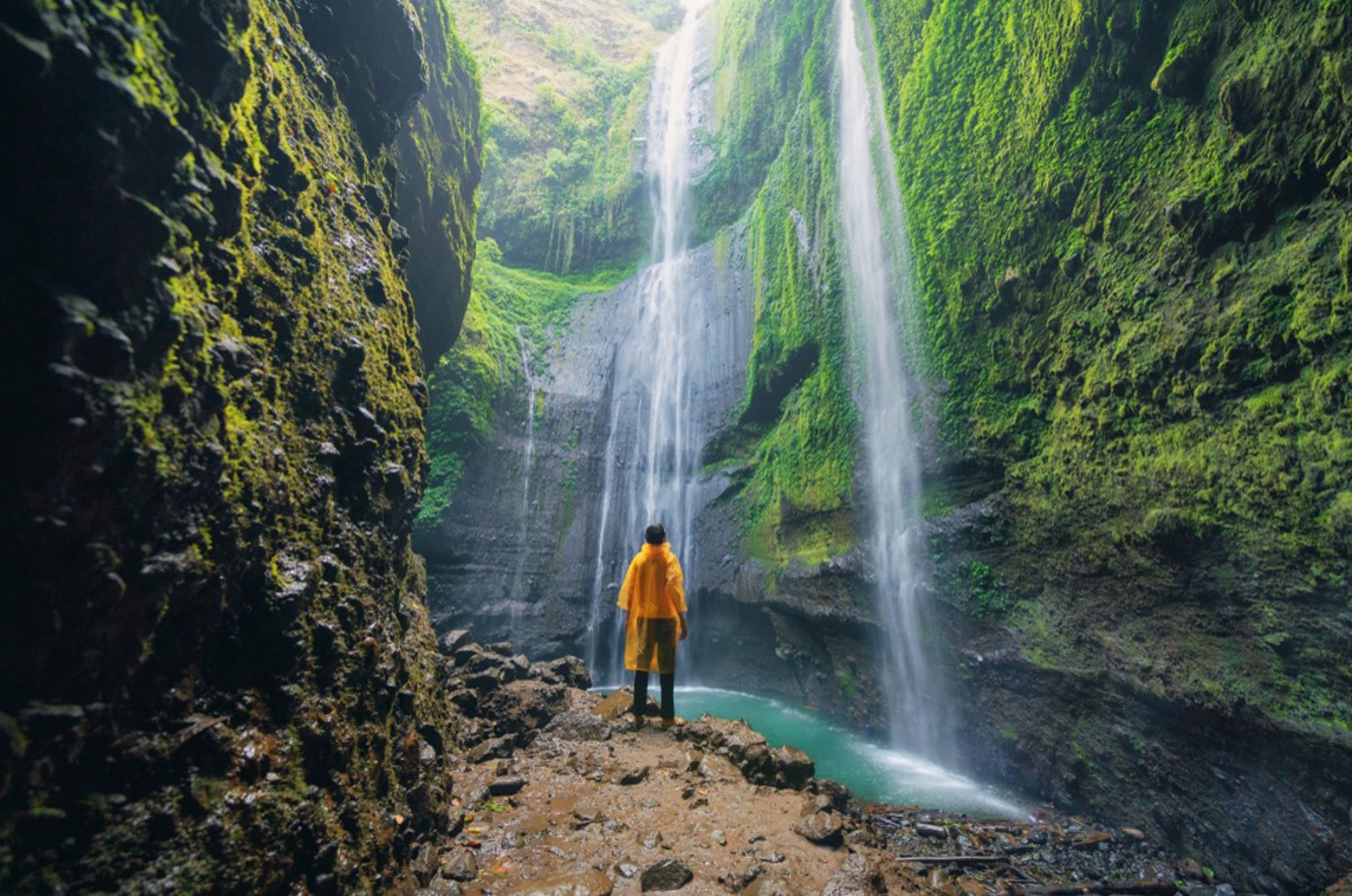 Bezoeker in gele regenjas bij een hoge waterval in een groene kloof in Indonesië.