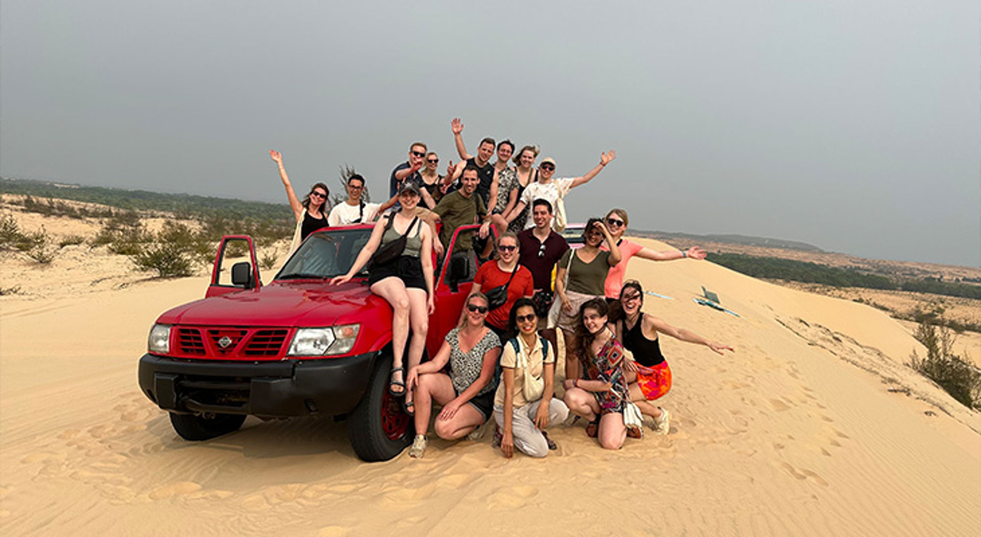 Jeeptour Mui Ne Groep reizigers poseert bij een rode 4x4 op zandduinen tijdens een woestijnexcursie.