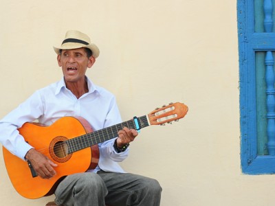 Cubaanse straatmuzikant met hoed zingt en speelt gitaar naast een blauw raam.