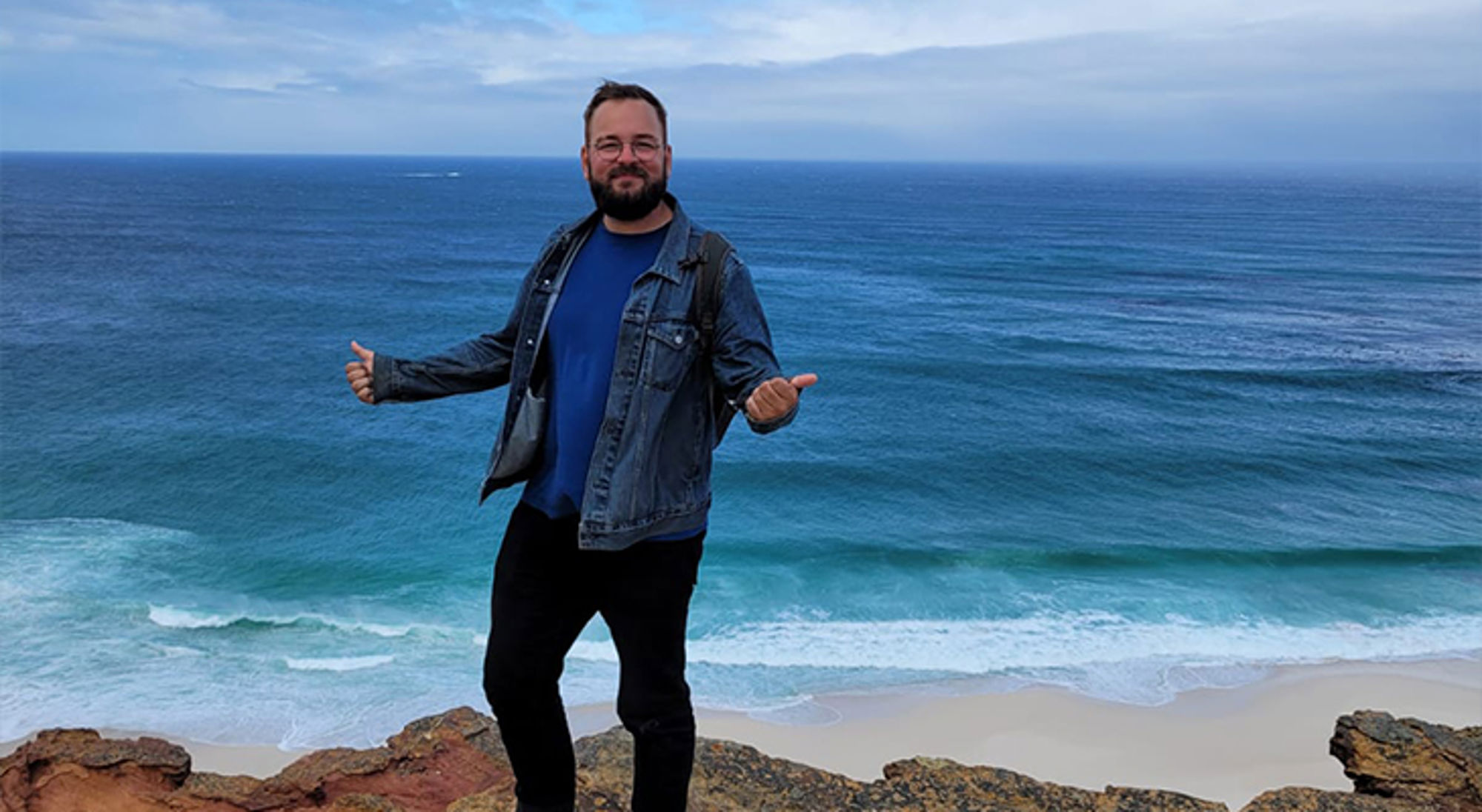 Ethan Man op een rots met uitgestrekte armen, met uitzicht op de oceaan en een zandstrand.